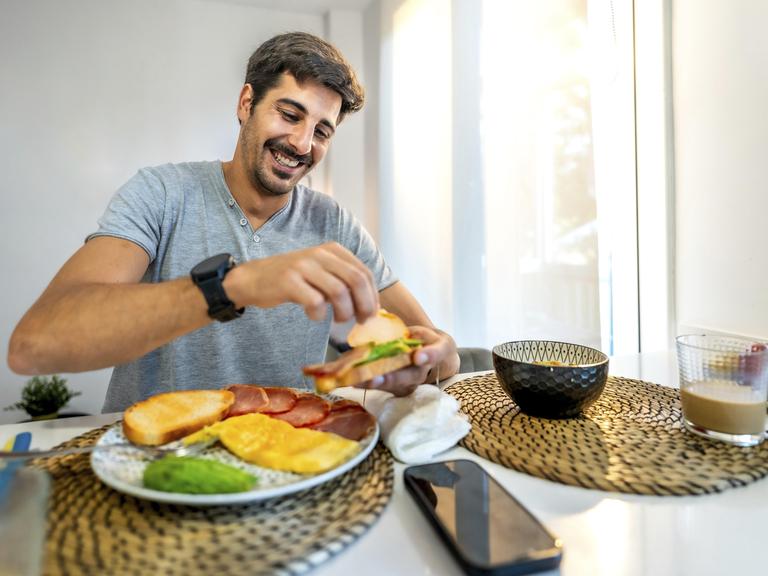 Ein Mann sitzt in der Küche am Frühstückstisch. Vor ihm stehen verschiedene Lebensmittel wie Obst, Brot, Müsli. Zu trinken hat er einen Milchkaffee. Der Mann belegt sich gerade ein Brot. Sein Handy liegt neben dem Teller. Ein Mann sitzt in der Küche am Frühstückstisch. Vor ihm stehen verschiedene Lebensmittel wie Obst, Brot, Müsli. Zu trinken hat er einen Milchkaffee. Der Mann belegt sich gerade ein Brot. Sein Handy liegt neben dem Teller.