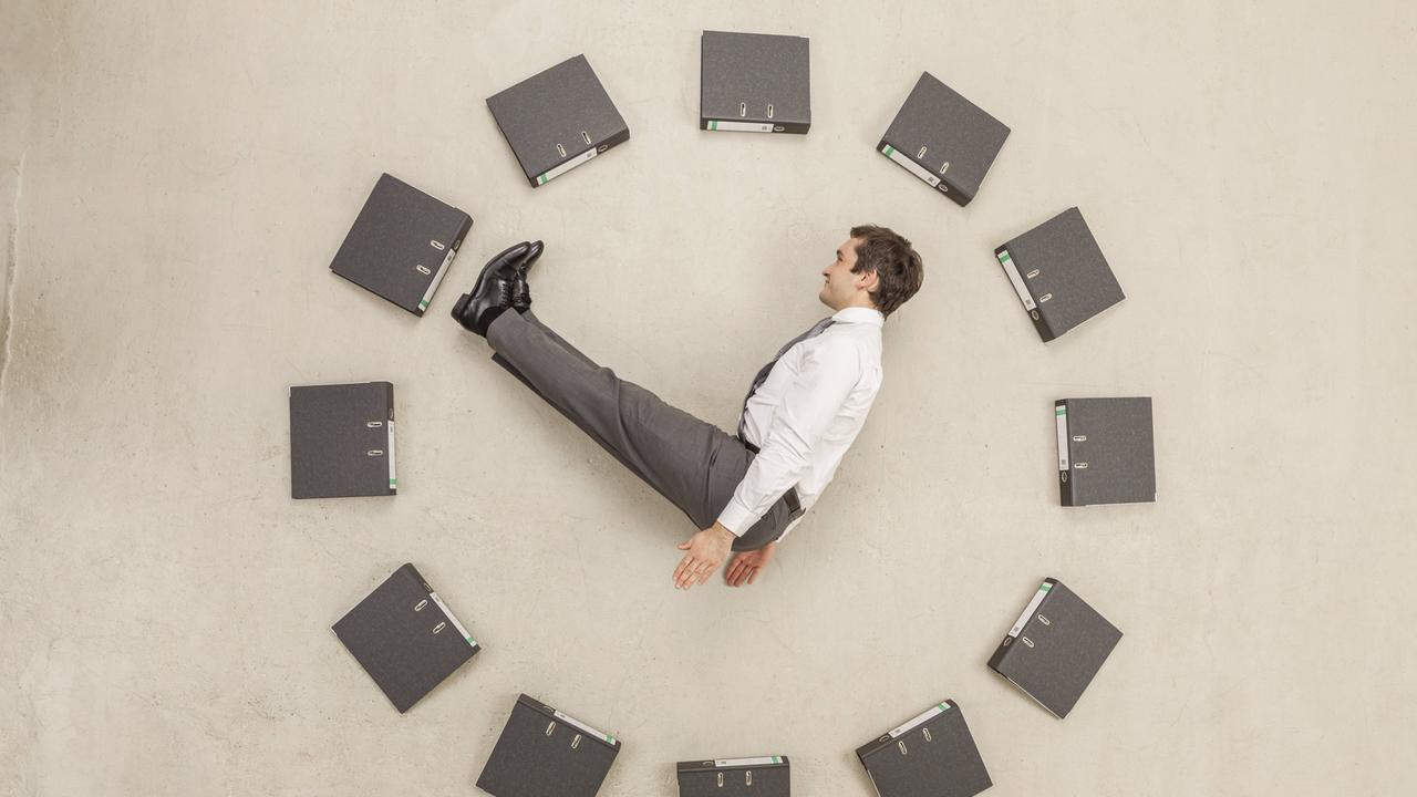 Aktenordner - kreisförmig auf dem Boden angeordnet - bilden eine Uhr. Ein Mann in Anzug stellt die Zeiger dar.