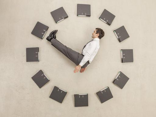 Aktenordner - kreisförmig auf dem Boden angeordnet - bilden eine Uhr. Ein Mann in Anzug stellt die Zeiger dar. Aktenordner - kreisförmig auf dem Boden angeordnet - bilden eine Uhr. Ein Mann in Anzug stellt die Zeiger dar.