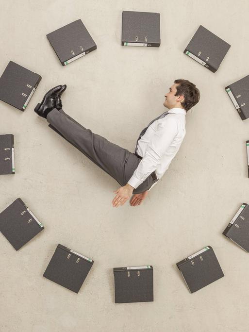 Aktenordner - kreisförmig auf dem Boden angeordnet - bilden eine Uhr. Ein Mann in Anzug stellt die Zeiger dar. Aktenordner - kreisförmig auf dem Boden angeordnet - bilden eine Uhr. Ein Mann in Anzug stellt die Zeiger dar.