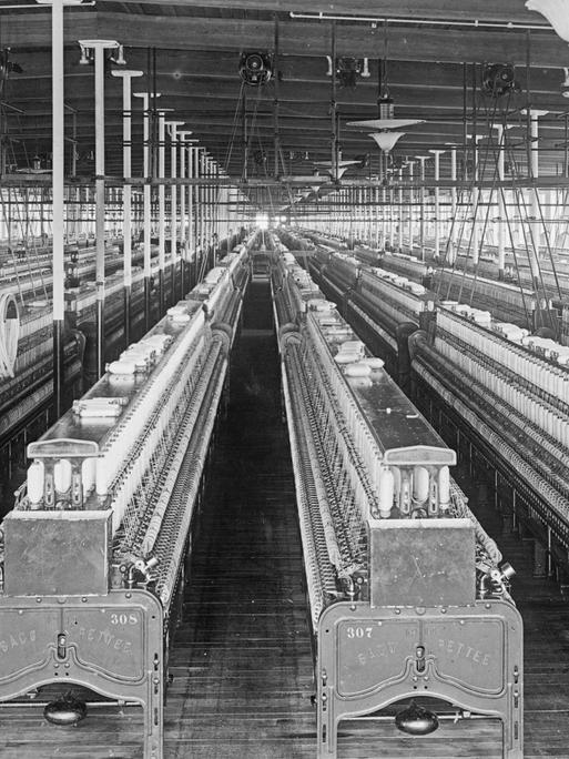 Schwarz-weiß-Foto: Blick in eine Baumwollspinnerei zwischen 1909 und 1920 in Manchester; eine große, menschenleere Fabrikhalle voller Webstühle.