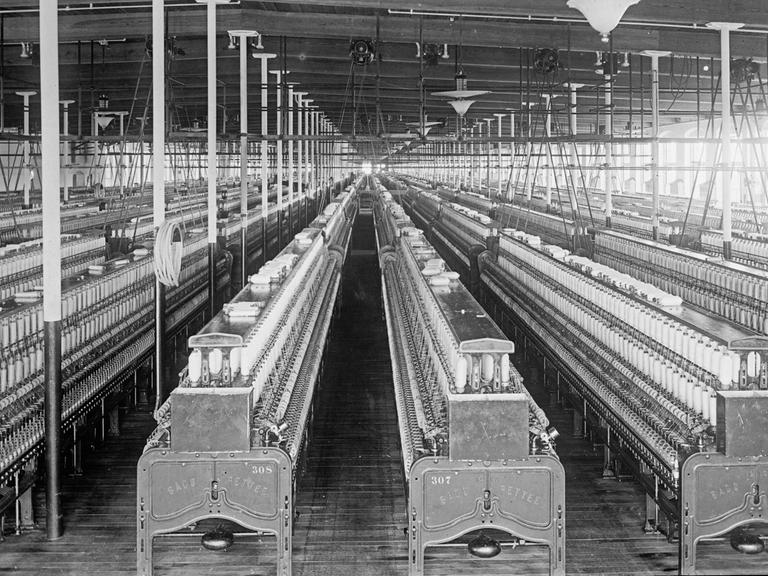 Schwarz-weiß-Foto: Blick in eine Baumwollspinnerei zwischen 1909 und 1920 in Manchester; eine große, menschenleere Fabrikhalle voller Webstühle.