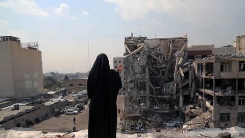 Das Bild zeigt eine verschleierte Person von hinten. Sie steht vor den Ruinen eines mehrstöckigen Gebäudes. 