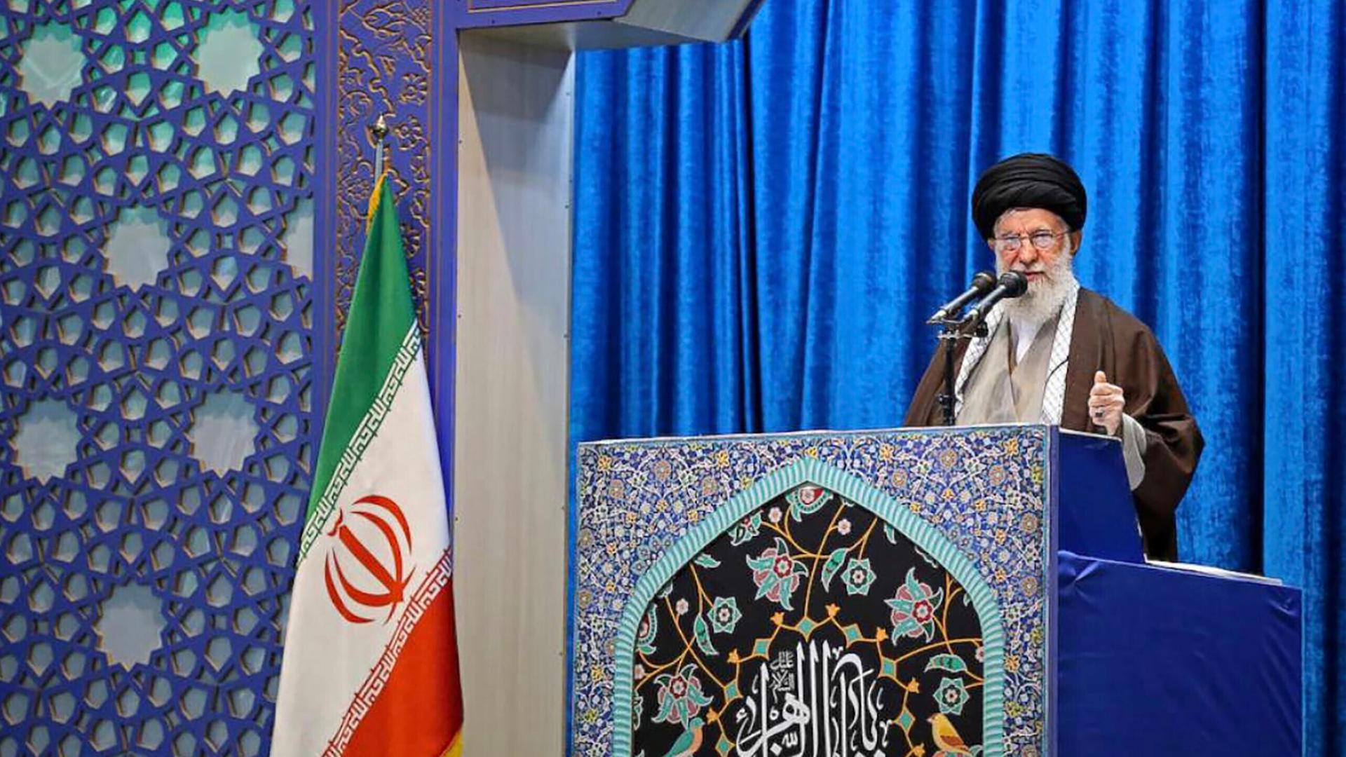 Ayatalloh Ali Khamenei bei Rede vor blauem Samthintergrund neben iranischer Flagge
