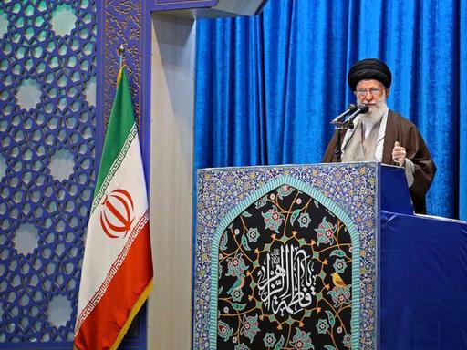 Ayatalloh Ali Khamenei bei Rede vor blauem Samthintergrund neben iranischer Flagge