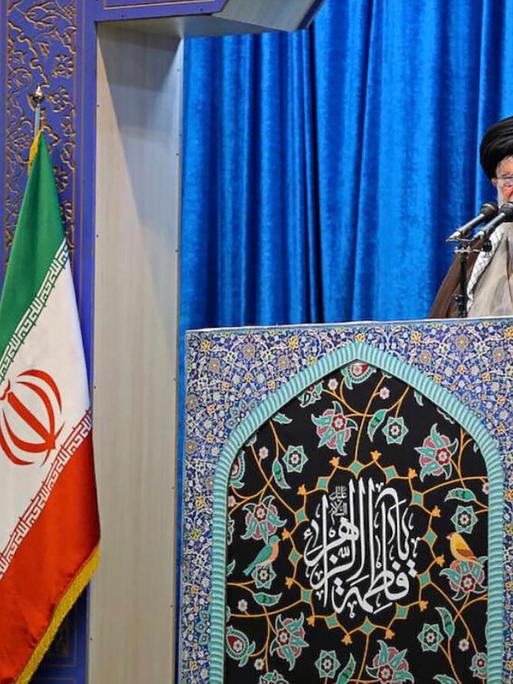 Ayatalloh Ali Khamenei bei Rede vor blauem Samthintergrund neben iranischer Flagge