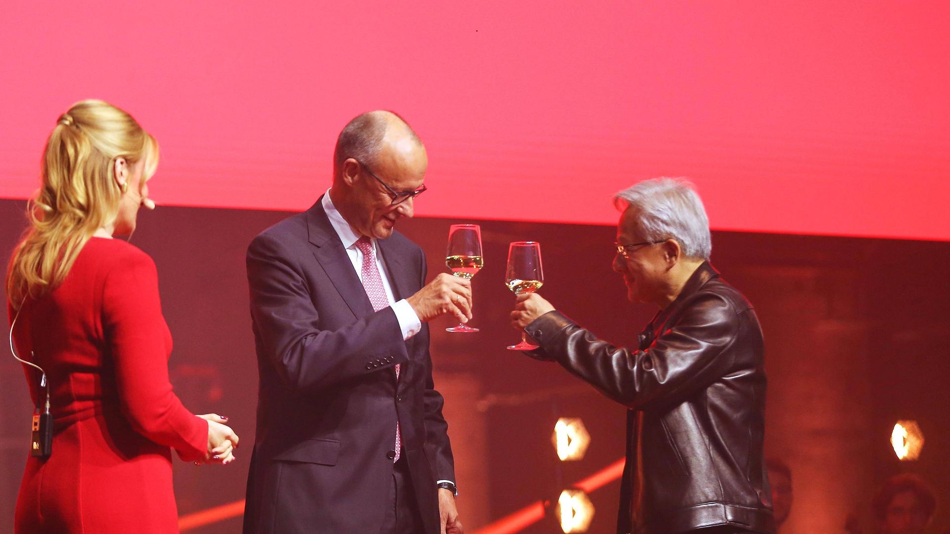 Bundeskanzler Friedrich Merz im dunklen Anzug und Nvidia-Chef Jensen Huang in seiner typischen schwarzen Lederjacke stoßen auf einer Bühne mit Weingläsern an. Links steht eine Moderatorin in einem roten Kleid mit dem Rücken zur Kamera, der Hintergrund… Bundeskanzler Friedrich Merz im dunklen Anzug und Nvidia-Chef Jensen Huang in seiner typischen schwarzen Lederjacke stoßen auf einer Bühne mit Weingläsern an. Links steht eine Moderatorin in einem roten Kleid mit dem Rücken zur Kamera, der Hintergrund…
