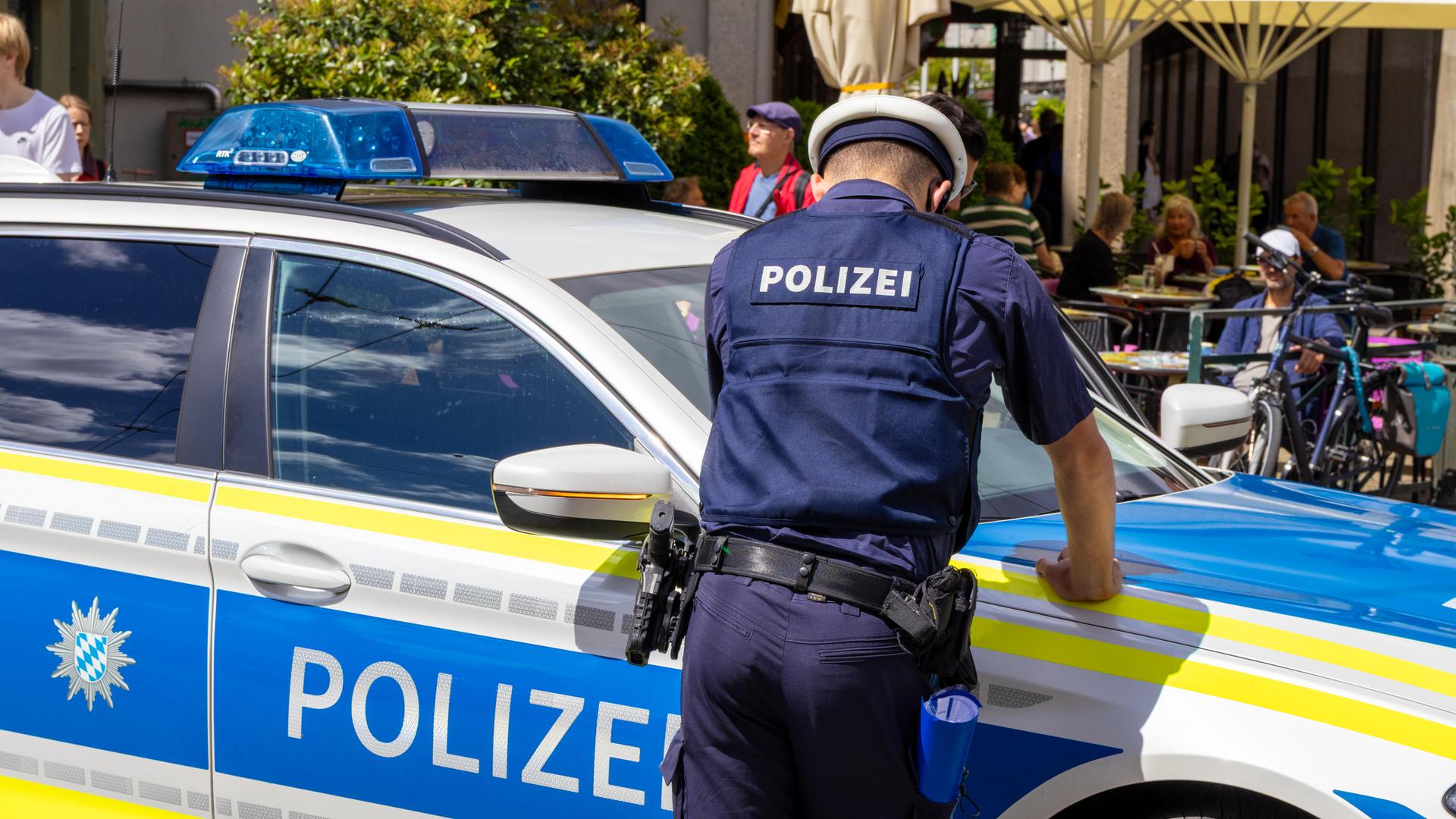 Ein Polizist lehnt an seinem Polizeiwagen in Augsburg. Er trägt Uniform. Im Hintergrund ist eine Fußgängerzone zu sehen.