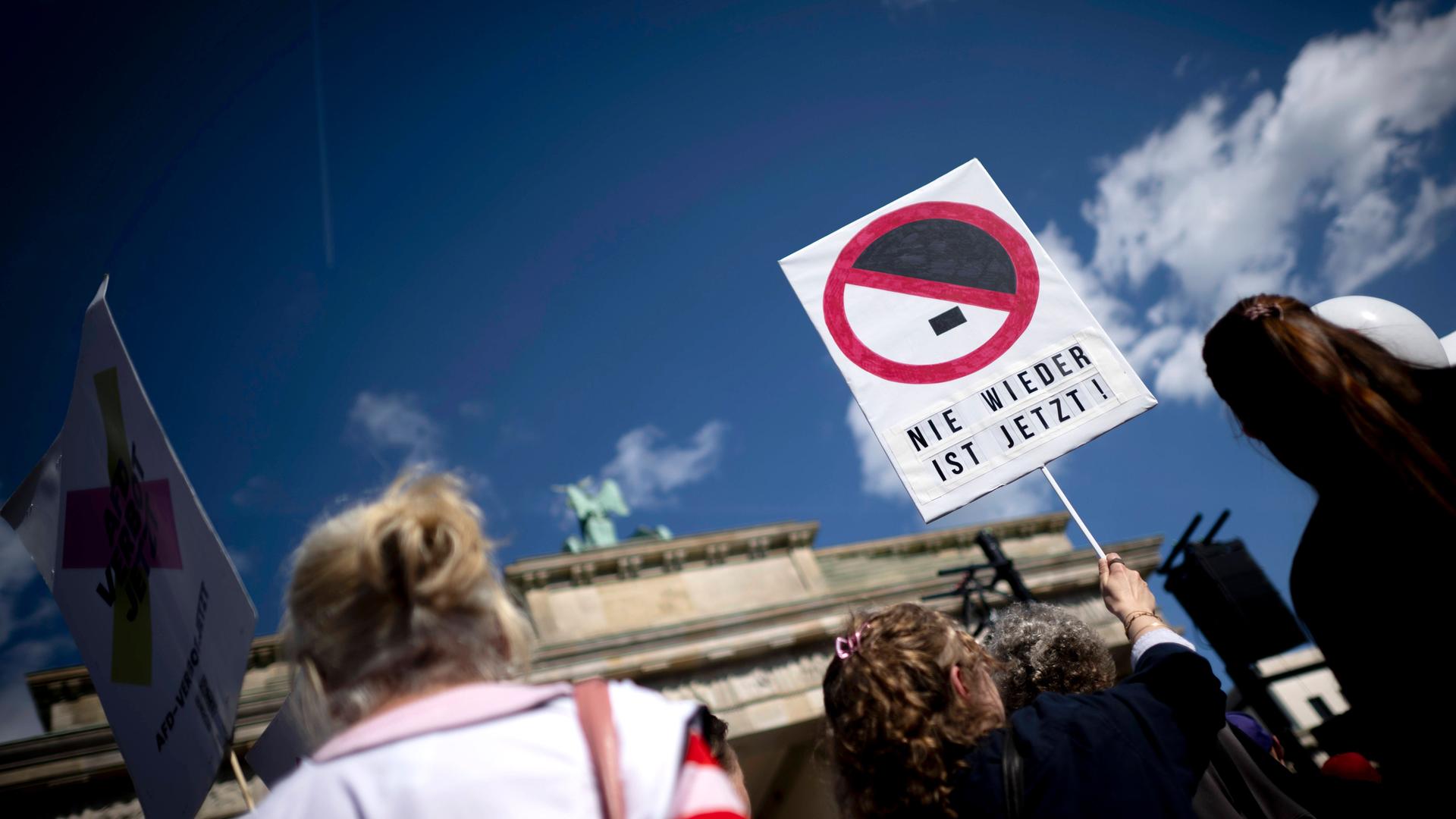 Demonstration gegen Rechtsextremismus vor dem Brandenburger Tor in Berlin; im Fokus hält eine Person ein Schild mit der Aufschrift „NIE WIEDER IST JETZT!“ unter einem durchgestrichenen Hitler-Schnitt. Demonstration gegen Rechtsextremismus vor dem Brandenburger Tor in Berlin; im Fokus hält eine Person ein Schild mit der Aufschrift „NIE WIEDER IST JETZT!“ unter einem durchgestrichenen Hitler-Schnitt.