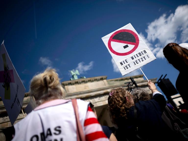 Demonstration gegen Rechtsextremismus vor dem Brandenburger Tor in Berlin; im Fokus hält eine Person ein Schild mit der Aufschrift „NIE WIEDER IST JETZT!“ unter einem durchgestrichenen Hitler-Schnitt. Demonstration gegen Rechtsextremismus vor dem Brandenburger Tor in Berlin; im Fokus hält eine Person ein Schild mit der Aufschrift „NIE WIEDER IST JETZT!“ unter einem durchgestrichenen Hitler-Schnitt.