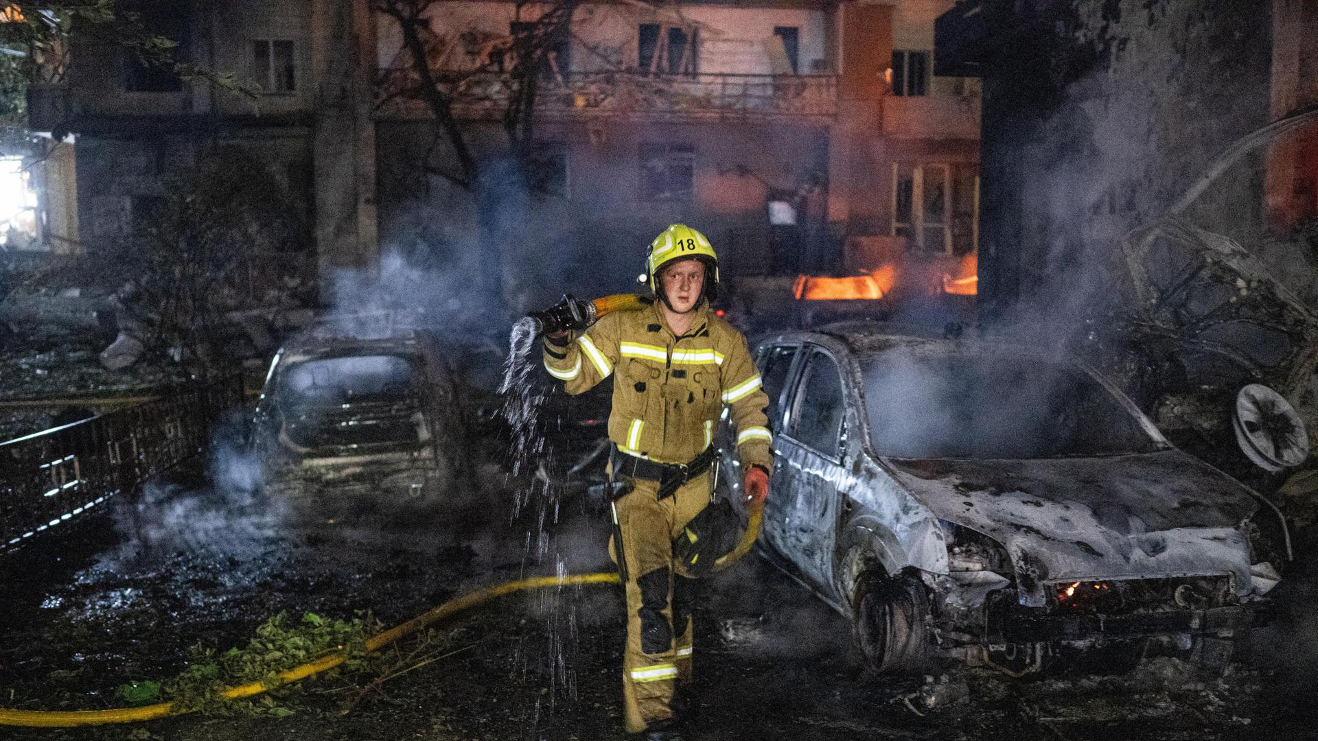 Nach einem russischen Raketenangriff in Kiew versucht eine Feuerwehrmann Brandherde zu löschen. Neben ihm stehen ausgebrannte Autos. Im Hintergrund lodern Feuer in einem Wohnhaus. Nach einem russischen Raketenangriff in Kiew versucht eine Feuerwehrmann Brandherde zu löschen. Neben ihm stehen ausgebrannte Autos. Im Hintergrund lodern Feuer in einem Wohnhaus.