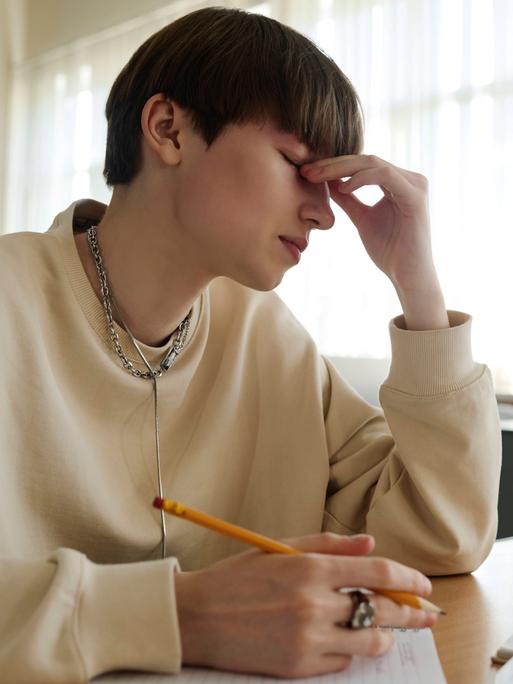 Eine Person sitzt in einer Bibliothek an einem Schreibtisch und hält einen Stift in der Hand. Mit der anderen Hand hält sich die Person an der Nase fest. Die Augen sind geschlossen.