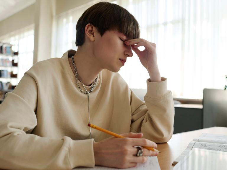 Eine Person sitzt in einer Bibliothek an einem Schreibtisch und hält einen Stift in der Hand. Mit der anderen Hand hält sich die Person an der Nase fest. Die Augen sind geschlossen.