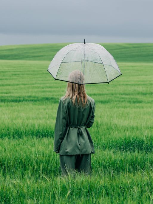 Modellfoto einer Frau, die mit einem durchsichtigen Regenschirm im Regen in einem grünen Weizenfeld steht Modellfoto einer Frau, die mit einem durchsichtigen Regenschirm im Regen in einem grünen Weizenfeld steht