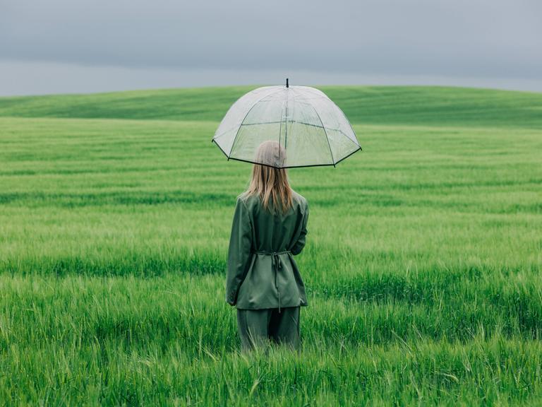Modellfoto einer Frau, die mit einem durchsichtigen Regenschirm im Regen in einem grünen Weizenfeld steht Modellfoto einer Frau, die mit einem durchsichtigen Regenschirm im Regen in einem grünen Weizenfeld steht