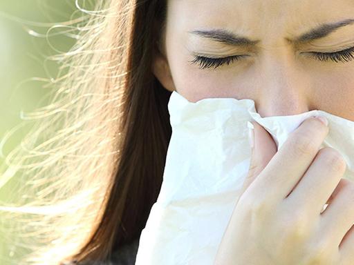 Eine Frau niest in ein Taschentusch. Sie ist draußen, im Hintergrund ist es grün.