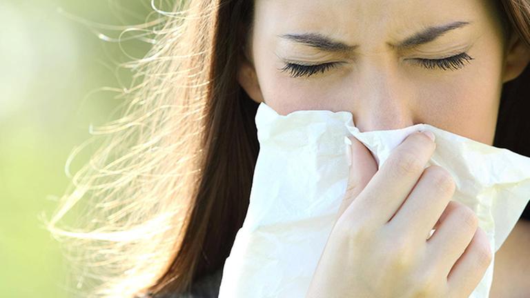 Eine Frau niest in ein Taschentusch. Sie ist draußen, im Hintergrund ist es grün.