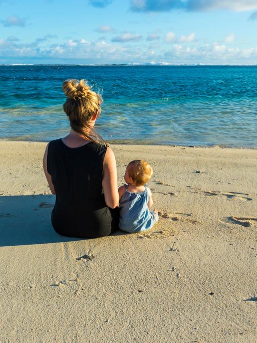 Eine Frau und ein Kind sitzen am Strand und schauen auf das Meer Eine Frau und ein Kind sitzen am Strand und schauen auf das Meer