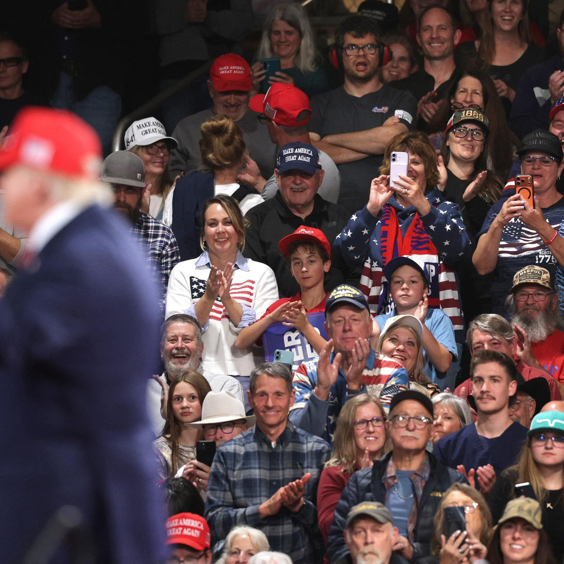 Anhänger sitzen bei einer Wahlkampfveranstaltung anläslich der Midterms in der Majestic Valley Arena in Kalispell; im Vordergrund steht US-Präsident Donald Trump zu sehen (14. Oktober 2026, Montana, USA)