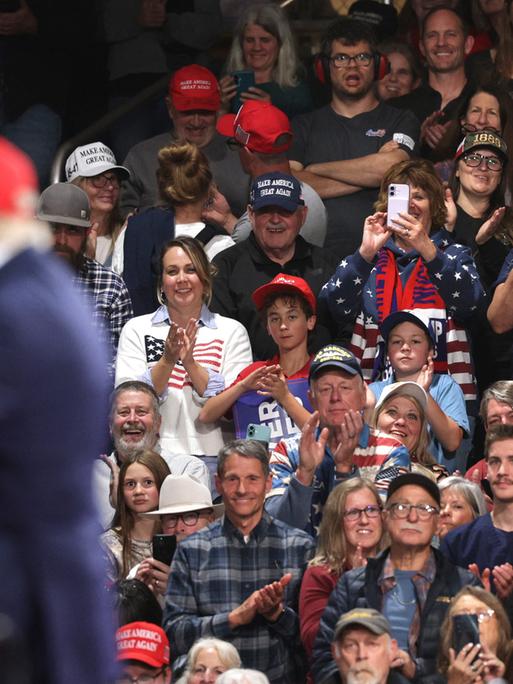 Anhänger sitzen bei einer Wahlkampfveranstaltung anläslich der Midterms in der Majestic Valley Arena in Kalispell; im Vordergrund steht US-Präsident Donald Trump zu sehen (14. Oktober 2026, Montana, USA)