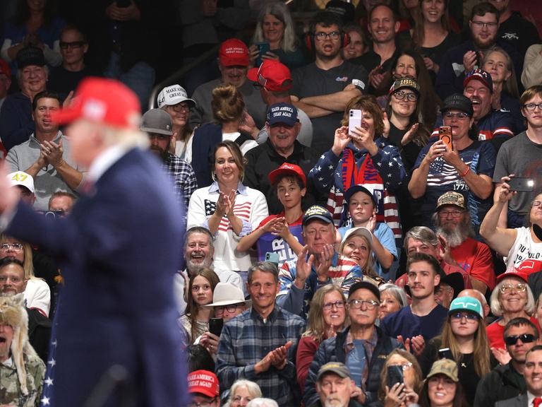 Anhänger sitzen bei einer Wahlkampfveranstaltung anläslich der Midterms in der Majestic Valley Arena in Kalispell; im Vordergrund steht US-Präsident Donald Trump zu sehen (14. Oktober 2026, Montana, USA)