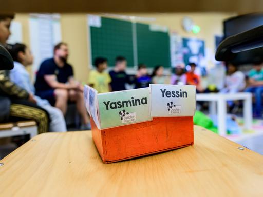 Schüler einer Grundschuleklasse sitzen vor der Tafel. Ebenso der Lehrer. Im Fotovordergrund steht auf einem Tisch eine Box mit zwei Namenskarten mit Yasmina und Yessin.