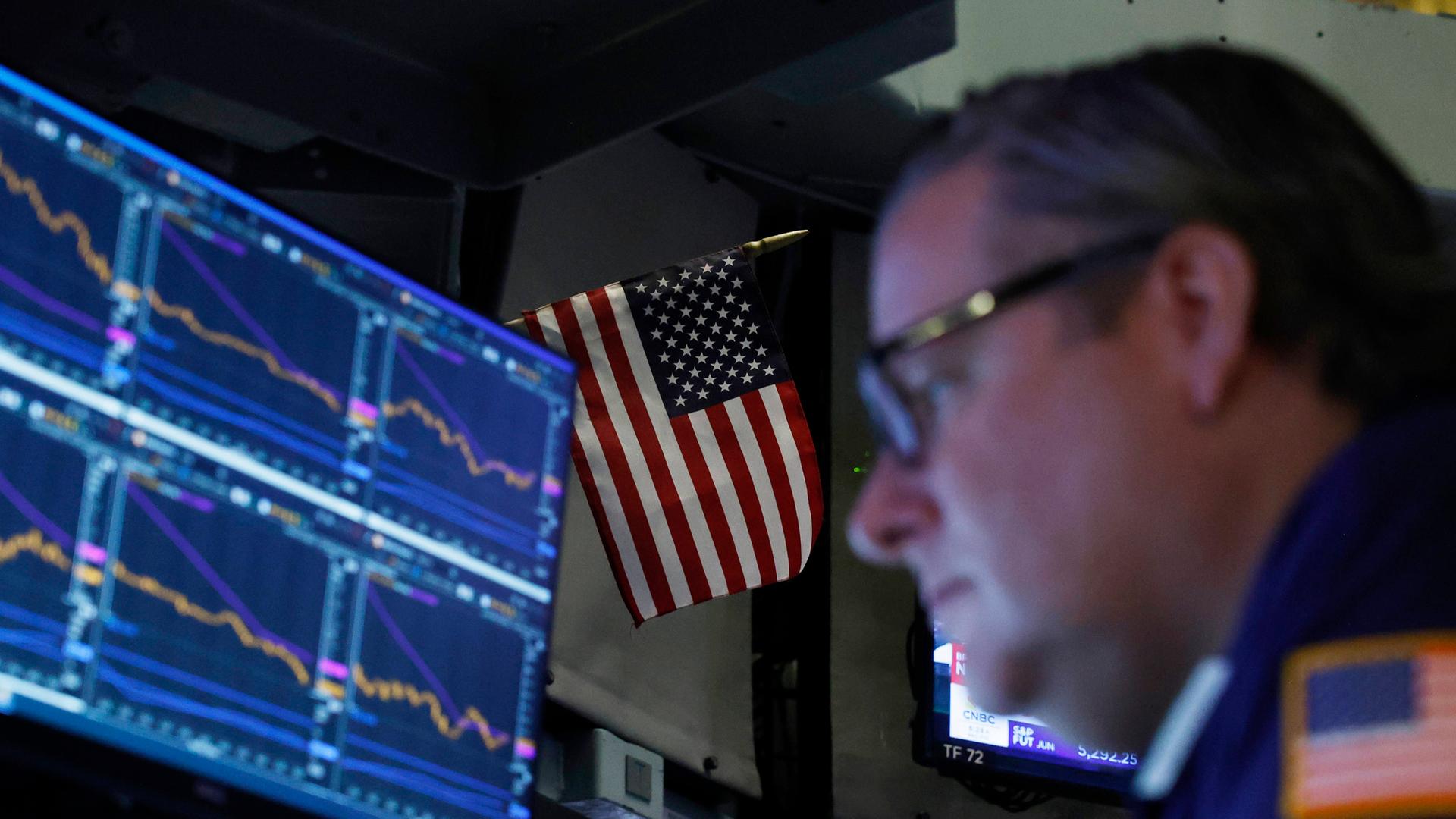 Ein Börsenhändler vor einem Bildschirm mit Aktienkursen, dahinter eine USA-Flagge Ein Börsenhändler vor einem Bildschirm mit Aktienkursen, dahinter eine USA-Flagge