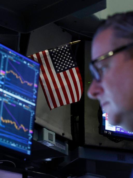 Ein Börsenhändler vor einem Bildschirm mit Aktienkursen, dahinter eine USA-Flagge Ein Börsenhändler vor einem Bildschirm mit Aktienkursen, dahinter eine USA-Flagge