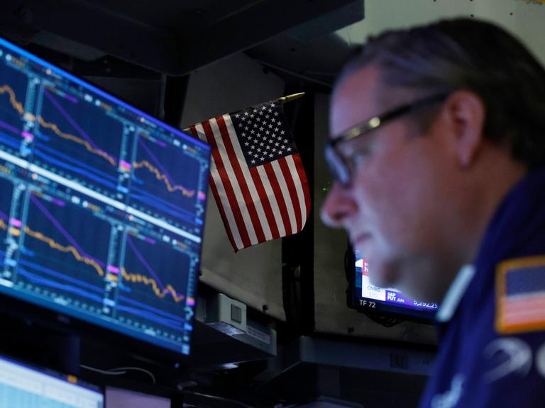 Ein Börsenhändler vor einem Bildschirm mit Aktienkursen, dahinter eine USA-Flagge Ein Börsenhändler vor einem Bildschirm mit Aktienkursen, dahinter eine USA-Flagge