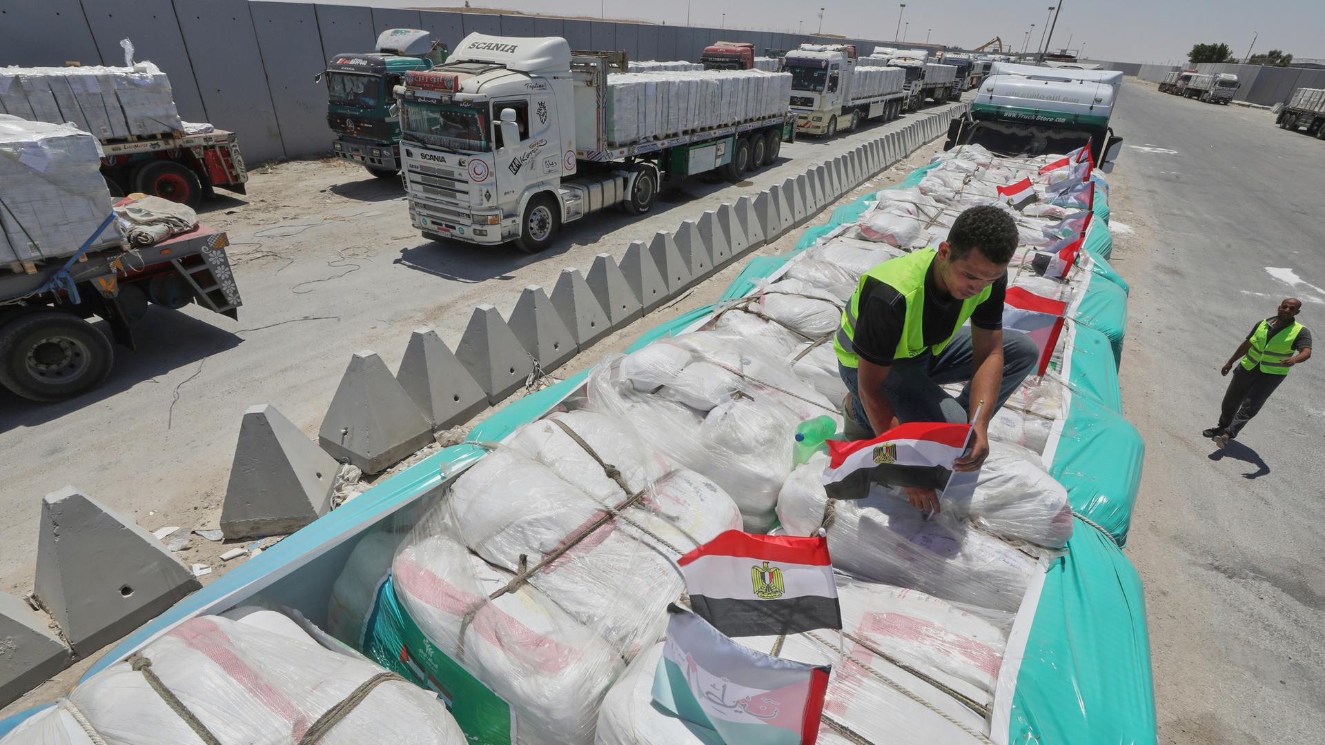 Mann mit gelber Warnweste befestigt ägyptische Flaggen auf Hilfslieferungen, die auf einem LKW liegen. Mehrere solcher LKWs stehen an der Grenze Gaza-Ägypten (06.08.2025)