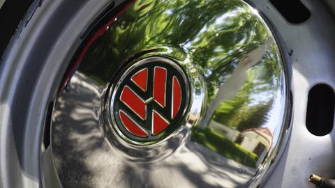 Nahaufnahme einer Pkw-Radkappe mit dem VW-Zeichen auf rotem Hintergrund. In der Radkappe spiegeln sich verschwommen eine Straße und ein Haus.