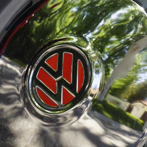 Nahaufnahme einer Pkw-Radkappe mit dem VW-Zeichen auf rotem Hintergrund. In der Radkappe spiegeln sich verschwommen eine Straße und ein Haus.