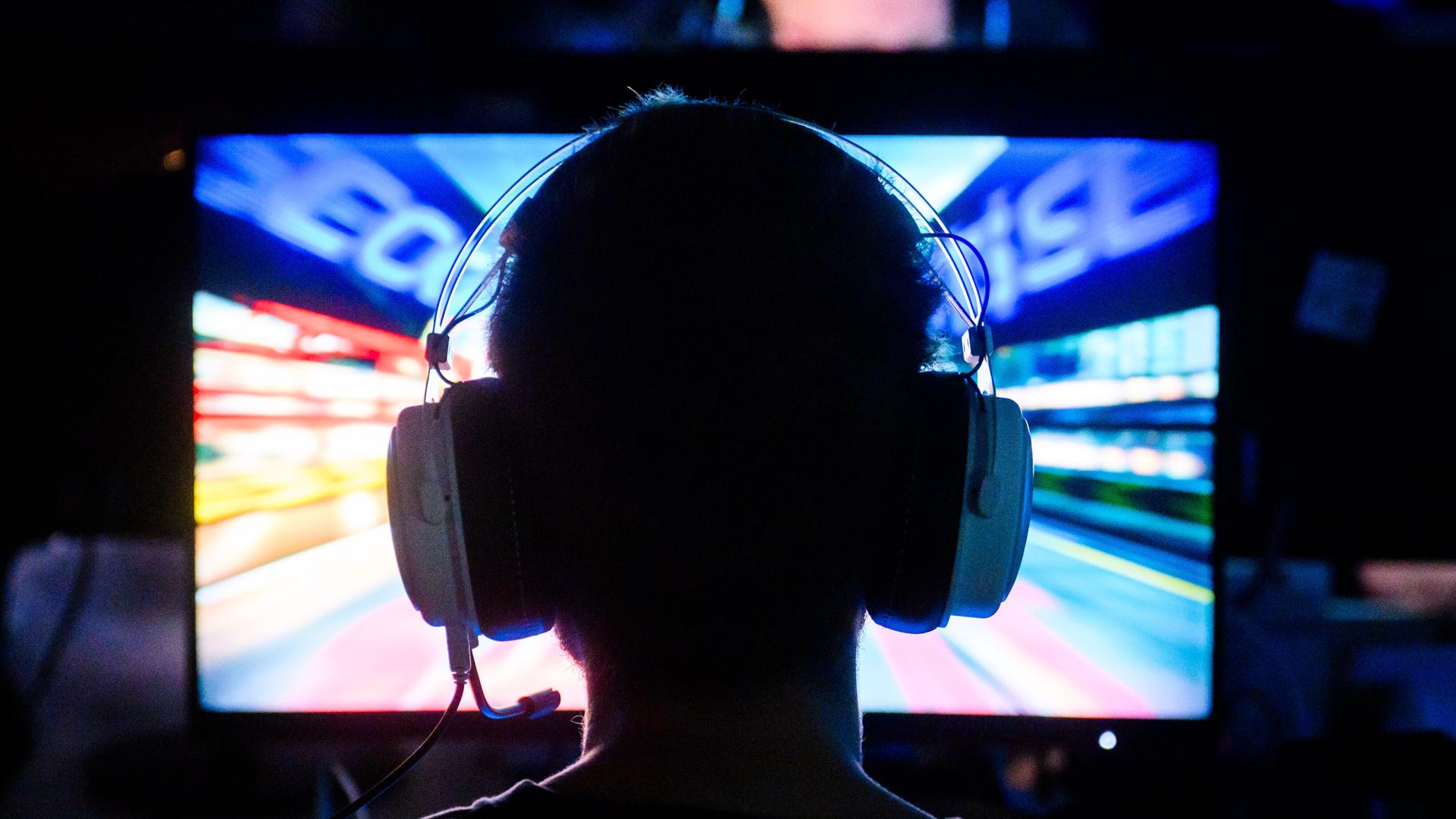 Ein Gamer spielt an ein Computerspiel. Wir sehen ihn von hinten. Er hat Kopfhörer mit Mikrofon auf.