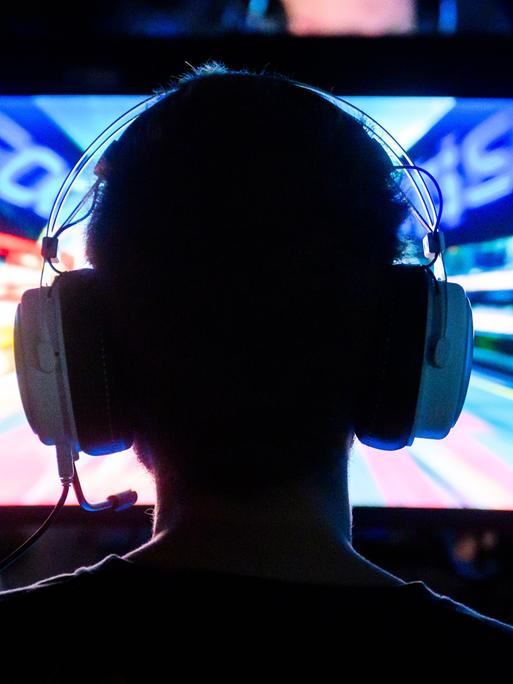 Ein Gamer spielt an ein Computerspiel. Wir sehen ihn von hinten. Er hat Kopfhörer mit Mikrofon auf.
