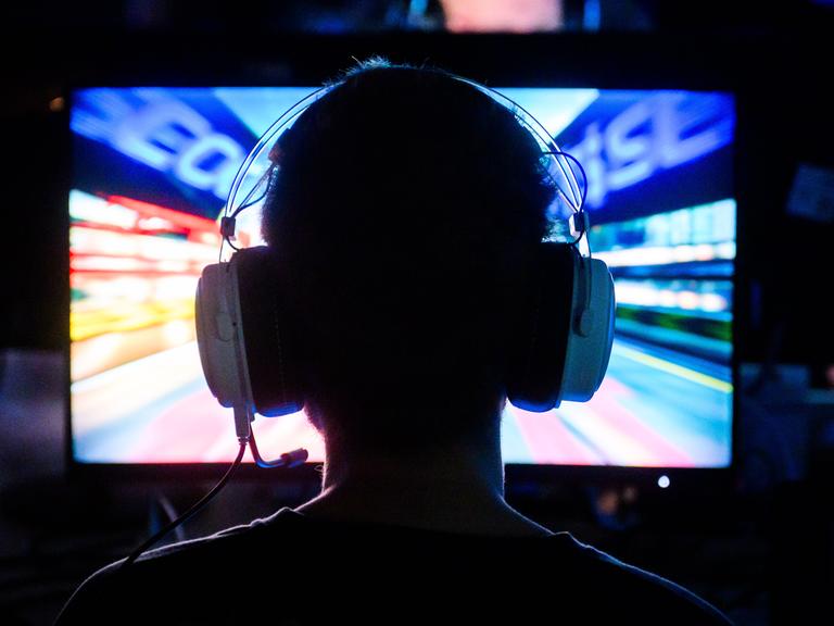 Ein Gamer spielt an ein Computerspiel. Wir sehen ihn von hinten. Er hat Kopfhörer mit Mikrofon auf.