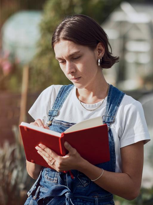 Eine Frau steht in einem Garten und schreibt etwas in ein rotes Notizbuch. Eine Frau steht in einem Garten und schreibt etwas in ein rotes Notizbuch.