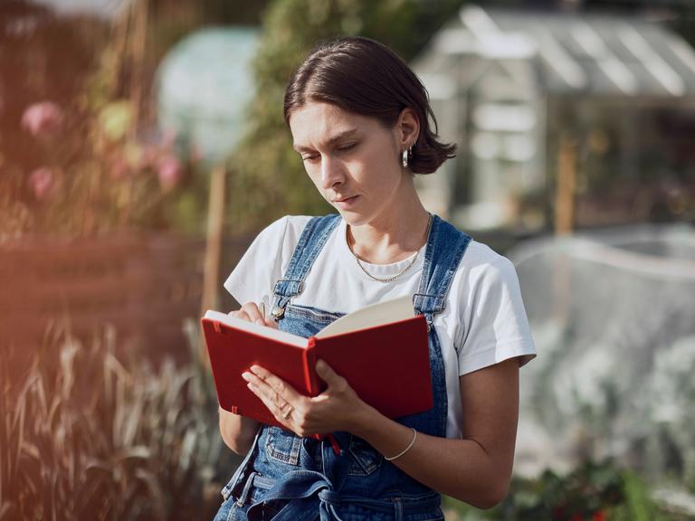 Eine Frau steht in einem Garten und schreibt etwas in ein rotes Notizbuch. Eine Frau steht in einem Garten und schreibt etwas in ein rotes Notizbuch.