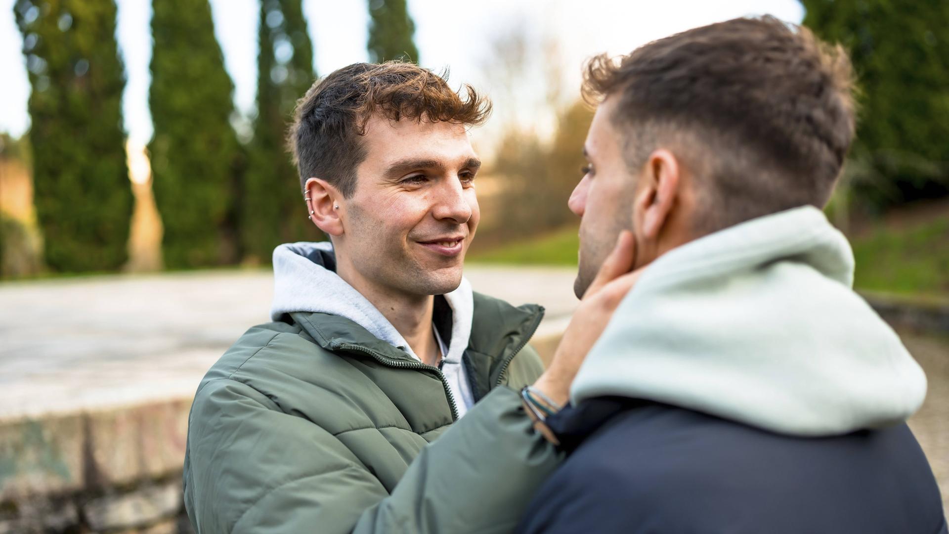 Zwei Männer stehen sich draußen gegenüber. Der eine Mann legt seine Hand an die Wange des anderen Mannes und lächelt. Beide tragen Winterjacken.
