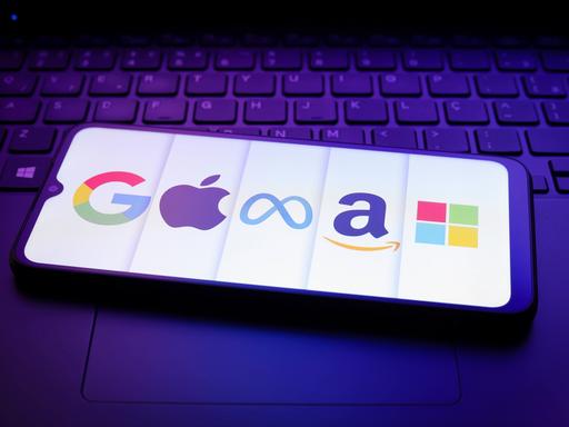 Die Logos von (linksnach rechts) Google, Apple, Meta, Amazon, Microsoft auf einem Smartphone-Display. Das Smartphone liegt auf der Tastatur eines Laptops