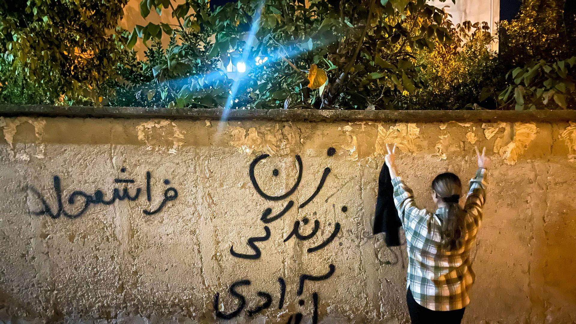 Eine junge Frau im Iran steht – ohne den obligatorischen Hijab – vor einer Mauer, mit dem Schriftzug: Frau, Leben, Freiheit.