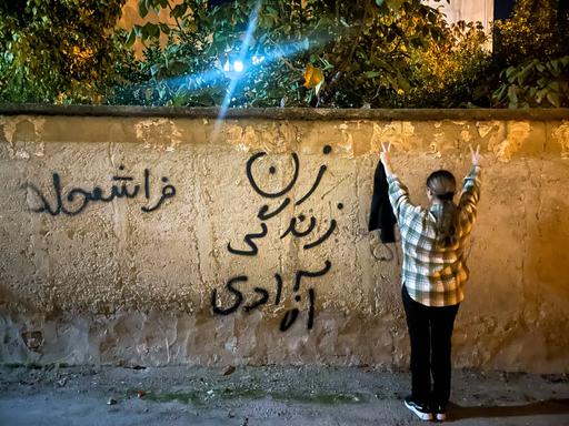 Eine junge Frau im Iran steht – ohne den obligatorischen Hijab – vor einer Mauer, mit dem Schriftzug: Frau, Leben, Freiheit.