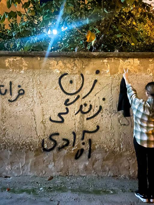 Eine junge Frau im Iran steht – ohne den obligatorischen Hijab – vor einer Mauer, mit dem Schriftzug: Frau, Leben, Freiheit.