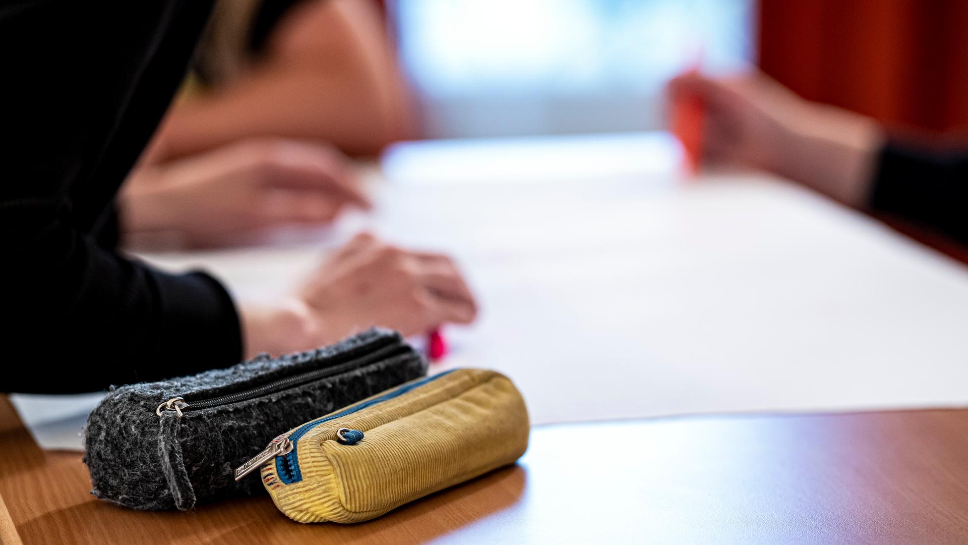 Zwei Federmäppchen liegen auf einem Tisch, im Hintergrund lehnt sich eine Person über den Tisch