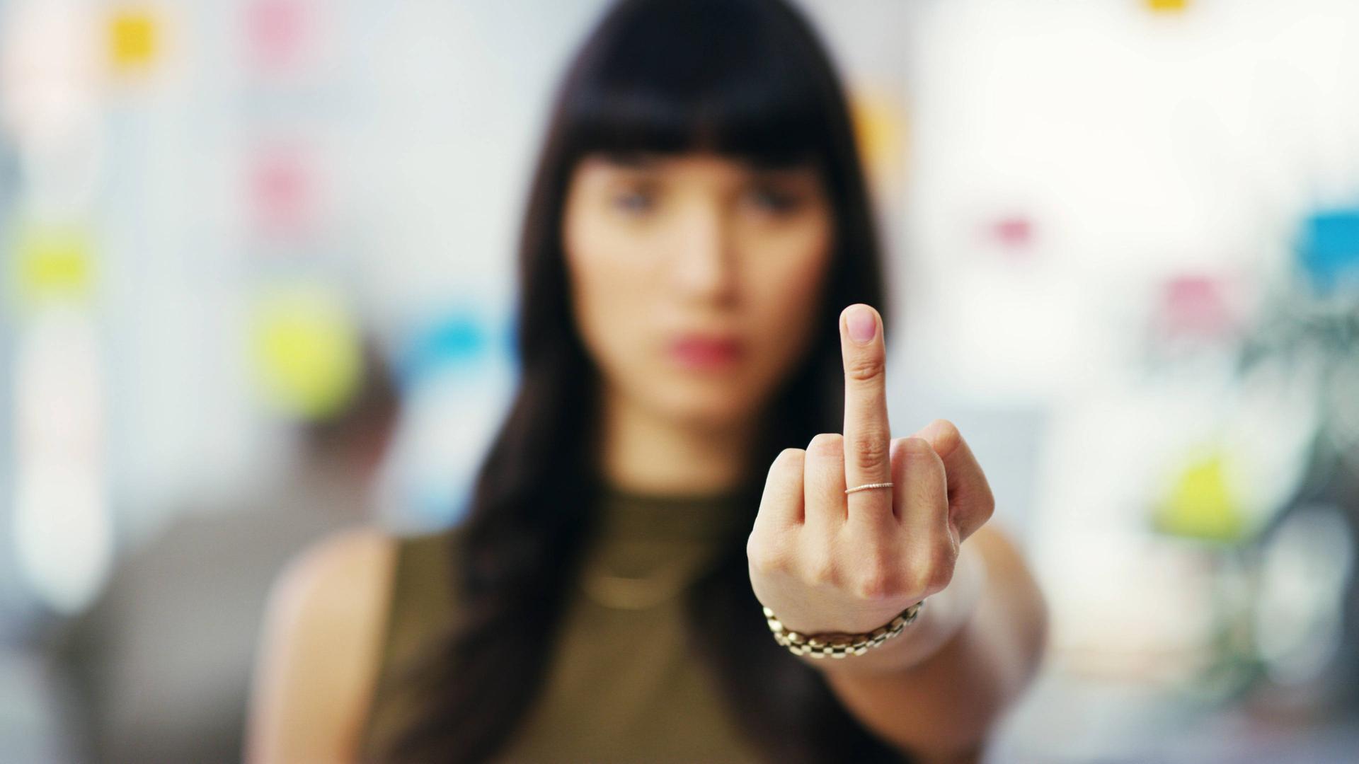 Foto einer jungen Frau in einem Büro, die eine Hand mit einem ausgestrecktem Stinkefinger in die Kamera hält und selbst im Hintergrund unscharf ist Foto einer jungen Frau in einem Büro, die eine Hand mit einem ausgestrecktem Stinkefinger in die Kamera hält und selbst im Hintergrund unscharf ist