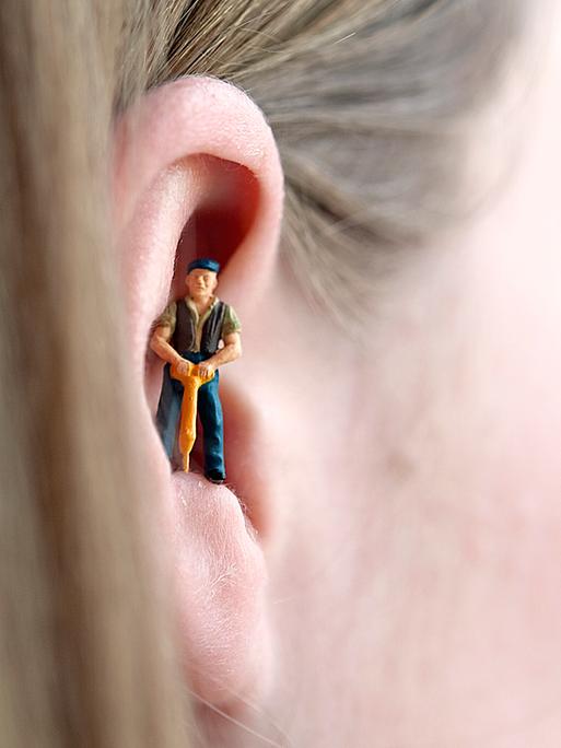 Eine Miniaturfigur eines Bauarbeiters mit Schlagbohrer steht im Ohr einer Frau