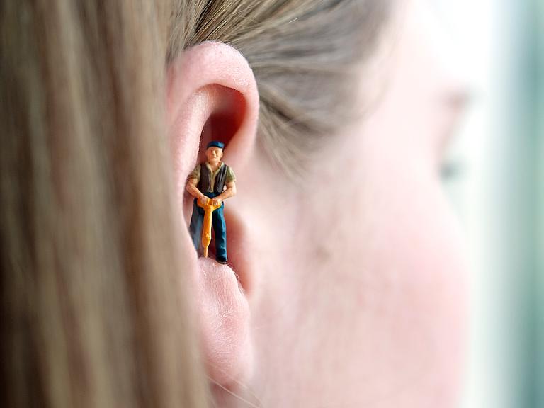 Eine Miniaturfigur eines Bauarbeiters mit Schlagbohrer steht im Ohr einer Frau