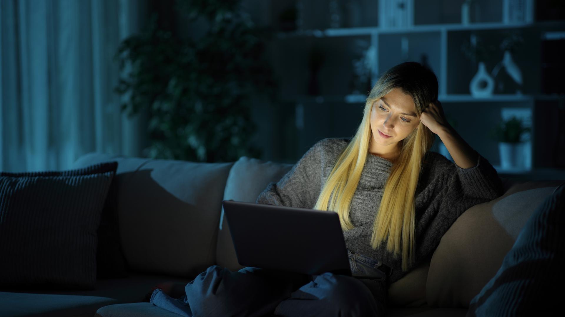 Eine Frau sitzt im Dunkeln auf einer Couch. Sie hat einen Laptop auf ihrem Schoß. Durch die Helligkeit des Bildschirms ist ihr Gesicht auch hell. Eine Frau sitzt im Dunkeln auf einer Couch. Sie hat einen Laptop auf ihrem Schoß. Durch die Helligkeit des Bildschirms ist ihr Gesicht auch hell.