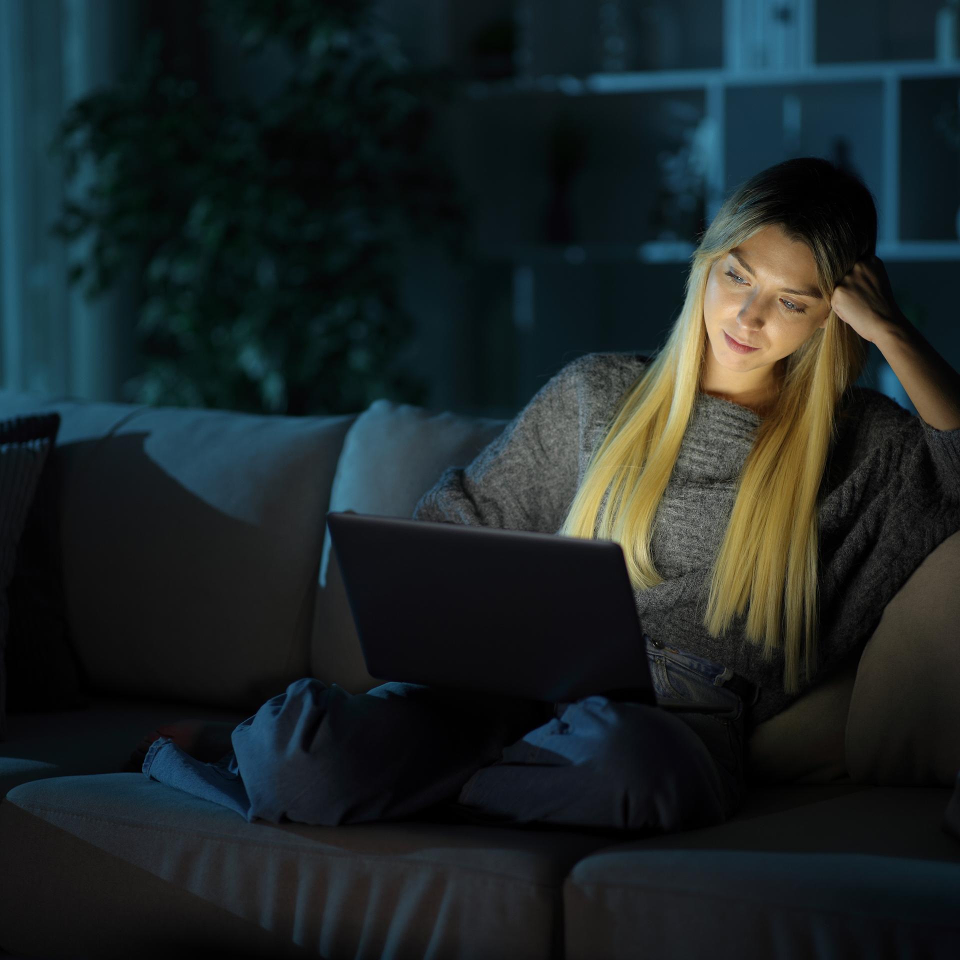 Eine Frau sitzt im Dunkeln auf einer Couch. Sie hat einen Laptop auf ihrem Schoß. Durch die Helligkeit des Bildschirms ist ihr Gesicht auch hell.