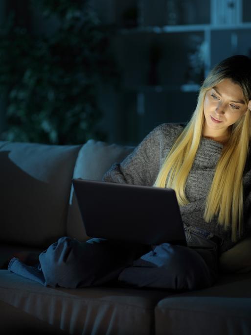 Eine Frau sitzt im Dunkeln auf einer Couch. Sie hat einen Laptop auf ihrem Schoß. Durch die Helligkeit des Bildschirms ist ihr Gesicht auch hell.