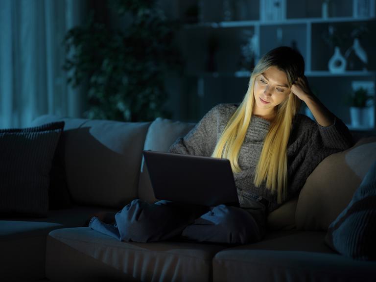 Eine Frau sitzt im Dunkeln auf einer Couch. Sie hat einen Laptop auf ihrem Schoß. Durch die Helligkeit des Bildschirms ist ihr Gesicht auch hell.